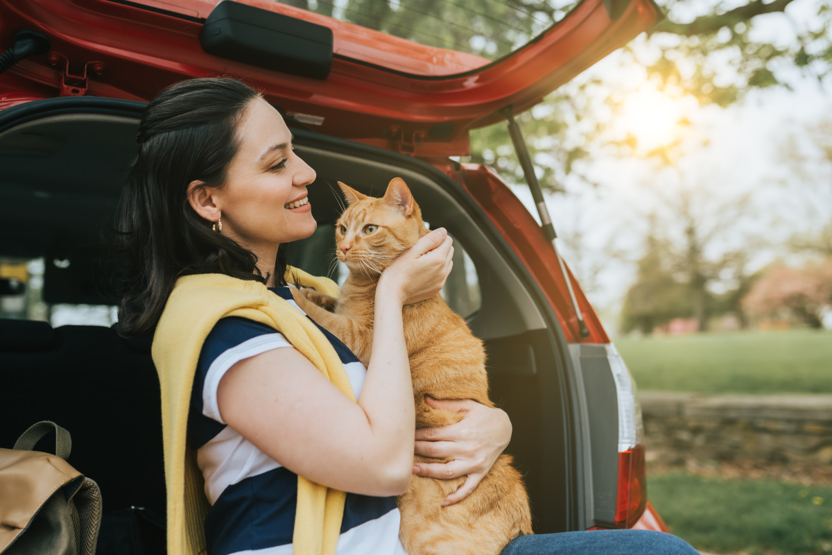 une femme souriante porte un chat roux dans ses bras devant le coffre ouvert de sa voiture rouge
